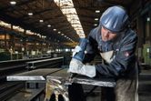 A person wearing safety equipment grinds metal with a straight grinder in a factory.