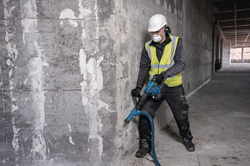 A person wearing safety equipment uses a demolition hammer on a concrete wall.
