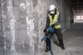 A person wearing safety equipment uses a demolition hammer on a concrete wall.