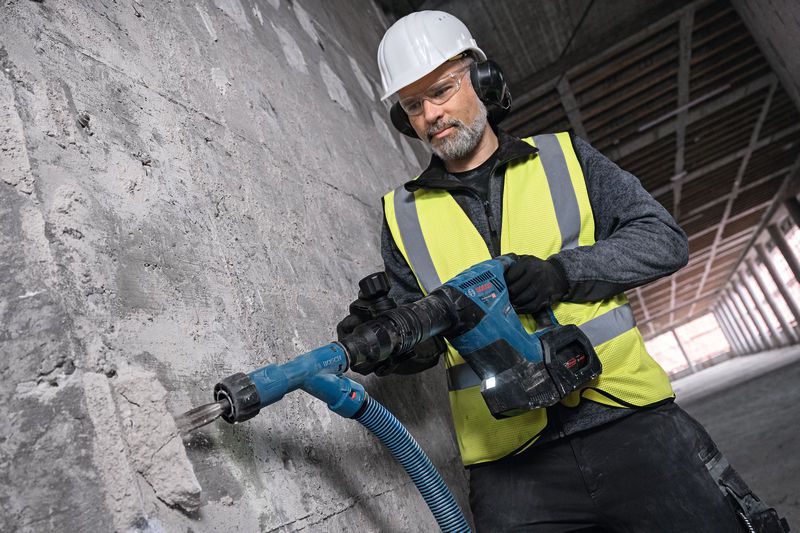 A worker wearing safety equipment drills into a concrete wall with a corded power tool.