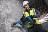 A worker wearing safety equipment drills into a concrete wall with a corded power tool.