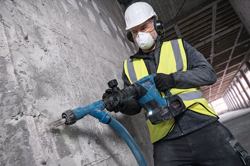 A worker wearing safety equipment drills into a concrete wall using a rotary hammer.