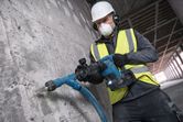 A worker wearing safety equipment drills into a concrete wall using a rotary hammer.