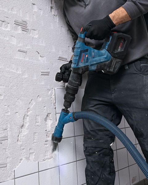 A person wearing safety equipment uses a rotary hammer drill with dust extraction on a tiled wall.