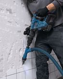 A person wearing safety equipment uses a rotary hammer drill with dust extraction on a tiled wall.