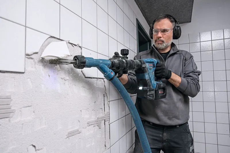 Person wearing safety equipment uses a power drill to remove wall tiles.