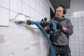 Person wearing safety equipment uses a power drill to remove wall tiles.