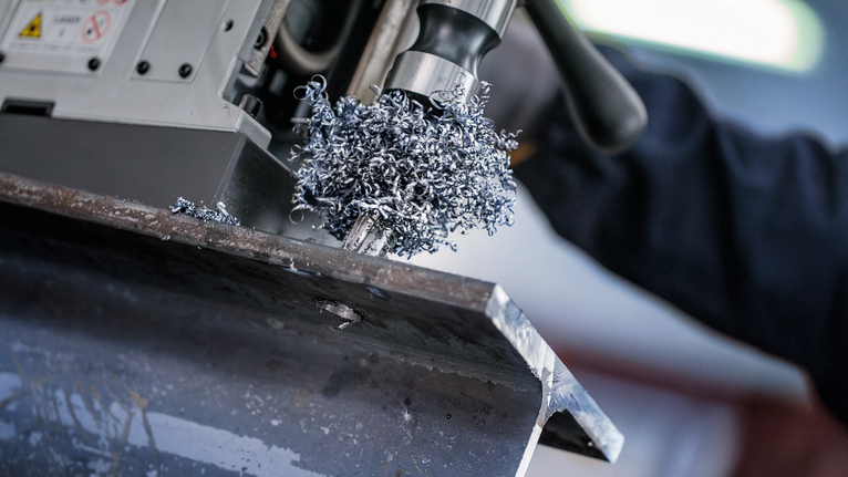 Person wearing safety equipment drills into a metal beam, creating metal shavings.