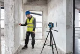 A person wearing safety equipment marks a wall guided by a laser leveling tool on a building tripod.
