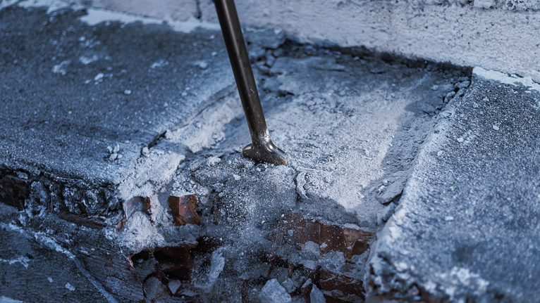 Chisel breaks through a concrete surface with dust and debris scattered.