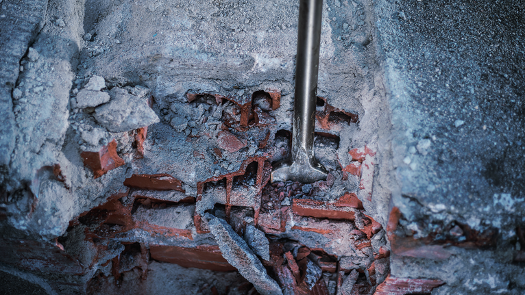 Rotary hammer chisel breaking through a damaged concrete and brick wall.