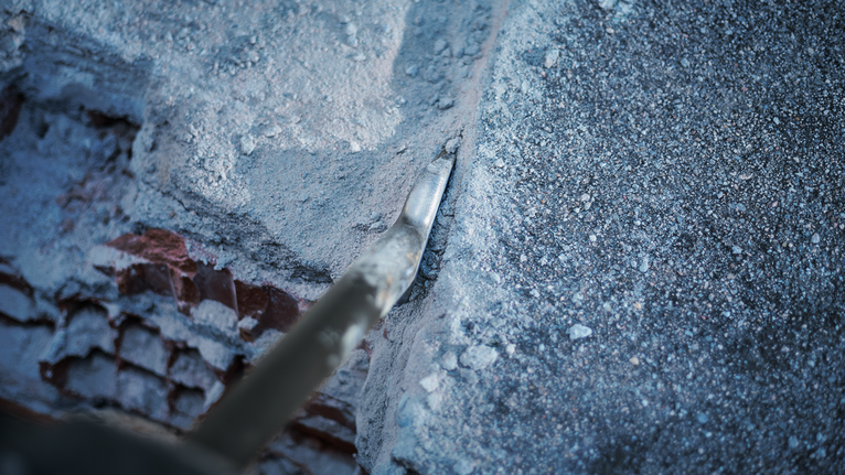 Pointed chisel breaking through concrete along a rough wall edge.