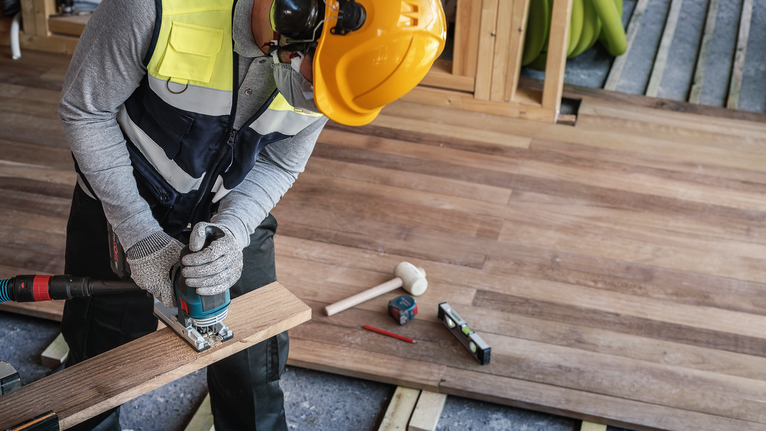 A person wearing safety equipment uses a jigsaw to cut wood flooring.