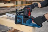 A person wearing safety equipment inserts a battery into a chainsaw placed on a wooden beam.