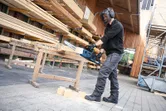 A person wearing safety equipment cuts wood outdoors with a cordless chainsaw.