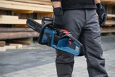 A person wearing safety equipment holds a cordless chainsaw at a lumber yard.