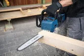 A person wearing safety equipment cuts a wooden beam using a blue chainsaw.