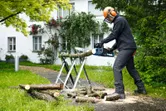 A person wearing safety equipment cuts wood logs outdoors with a cordless chainsaw.