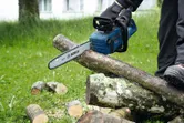 A person wearing safety equipment cuts branches outdoors using a chainsaw.