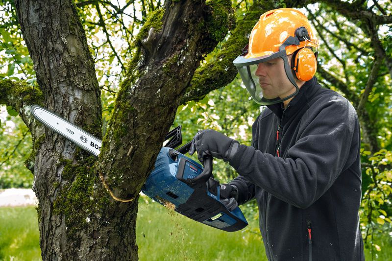A person wearing safety equipment cuts through a tree branch with a cordless chainsaw.
