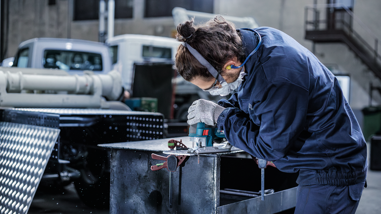 A person wearing safety equipment cuts sheet metal with a power tool in an industrial area.