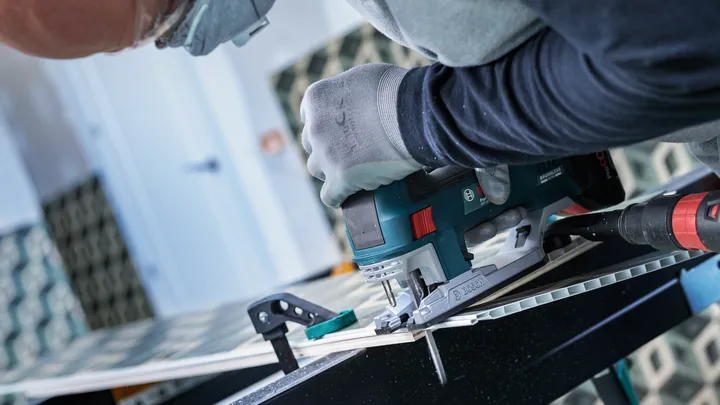 Person wearing safety equipment cuts tile with an electric jigsaw on a workbench.