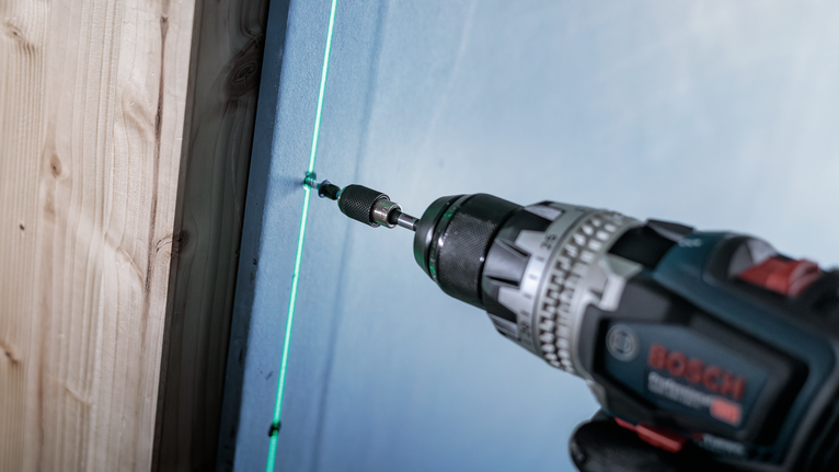 Cordless drill driving a screw into a wall along a green laser guide.