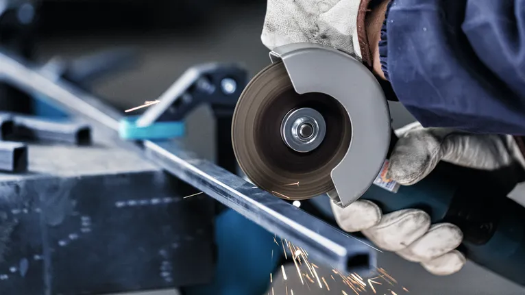 A worker wearing safety equipment cuts a metal bar with an angle grinder.