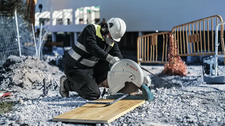 A person wearing safety equipment cuts a wooden plank with a circular saw at a construction site.