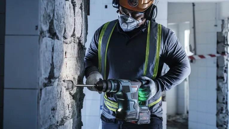 A person wearing safety equipment uses a rotary hammer to chisel a wall.