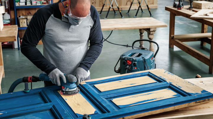 Object #200471068: 7390919_app_web_10 A worker wearing safety equipment sands an old wooden door in a workshop.