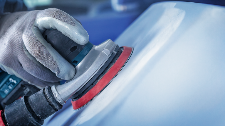 Person wearing safety equipment sanding a car surface with a power sander.