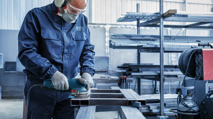 Object #200471078: 7390919_app_web_8 Worker wearing safety equipment grinds the surface of a metal beam in an industrial workshop.