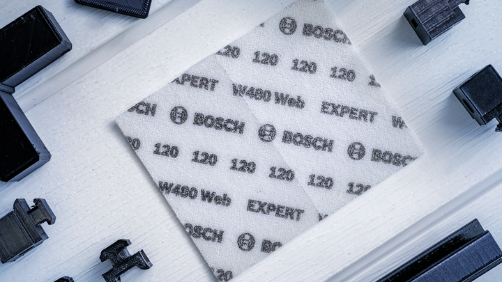 A square sanding sheet labeled with grit numbers lies on a white surface among black tool parts.