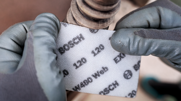 Person wearing safety equipment uses abrasive pad to sand a wooden surface.