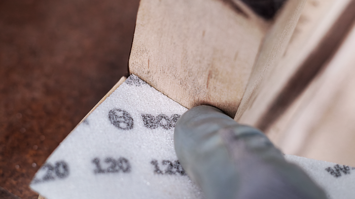 Gloved hand sanding a wooden edge with a foam abrasive sheet.