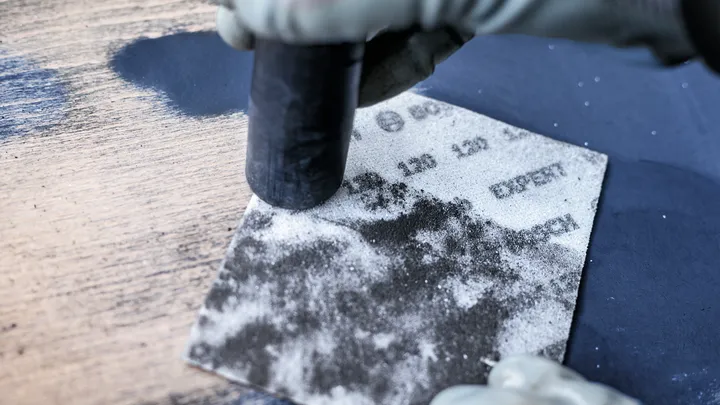 Gloved hand holds a sanding block over a dusty abrasive sheet.