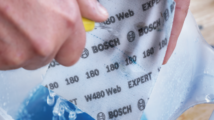 Person holding wet sandpaper sheet over a container filled with blue liquid.