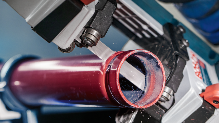 PRO CB2824 Band Saw Blade, 733 mm, 2 pcs A bandsaw slices through a red metal pipe in a workshop.