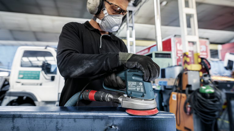 Person wearing safety equipment sands a metal surface with a power tool.