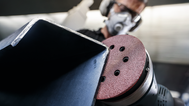 A person wearing safety equipment sands a metal beam with a power sander.