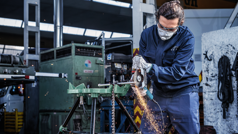 A person wearing safety equipment grinds metal, producing sparks in an industrial workshop.
