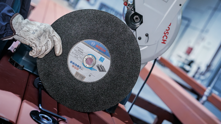 PRO Metal Cutting Disc, 305 x 3 x 25.4 mm Person wearing safety equipment holds a large abrasive cutoff wheel near a metal saw.