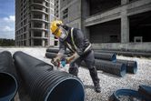 A person wearing safety equipment uses a cordless angle grinder to cut a large plastic pipe.