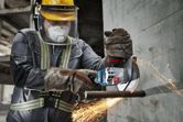 A person wearing safety equipment cuts rebar against a wall with a cordless angle grinder.
