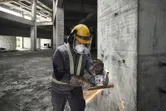 A person wearing safety equipment uses a cordless angle grinder to cut rebar on a concrete wall.