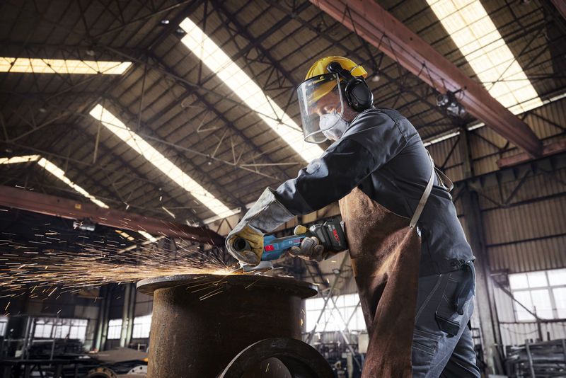 GWS 18V-15 S A person wearing safety equipment uses a cordless angle grinder to cut metal, creating sparks.