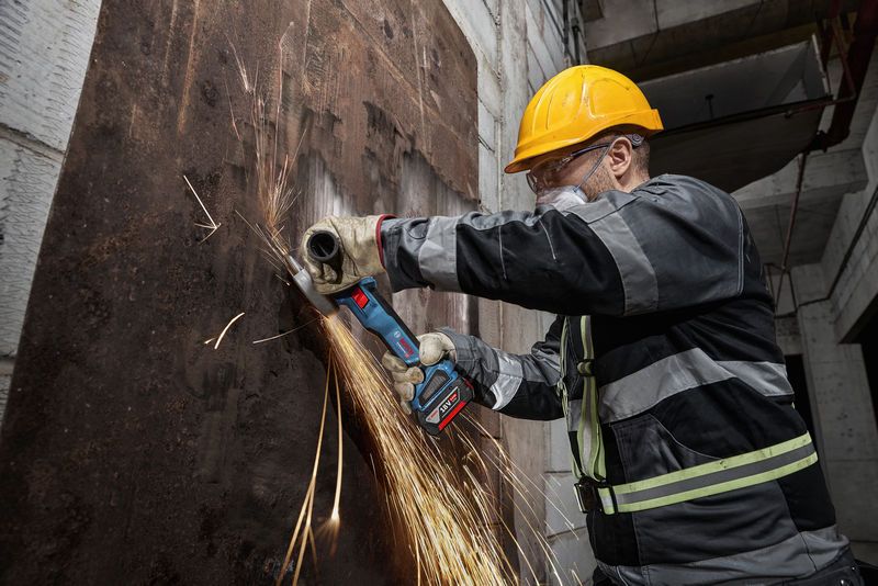 A person wearing safety equipment grinds a rusted metal sheet with a cordless angle grinder.