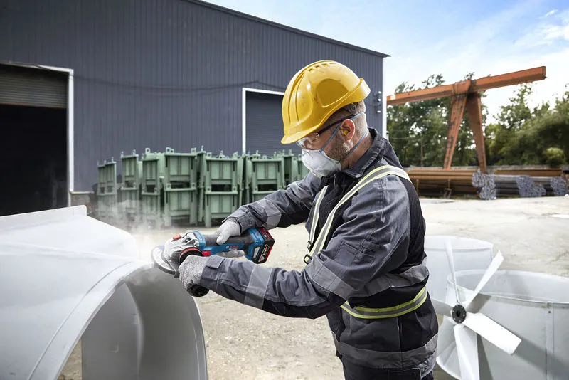 GWS 18V-11 S A person wearing safety equipment grinds metal outdoors at an industrial site.
