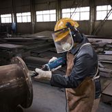 A person wearing safety equipment uses a straight grinder on a large metal pipe.